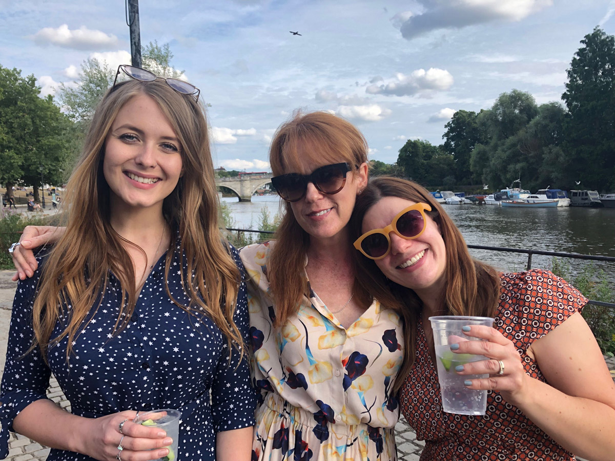 Three women looking to camera with drinks by a river